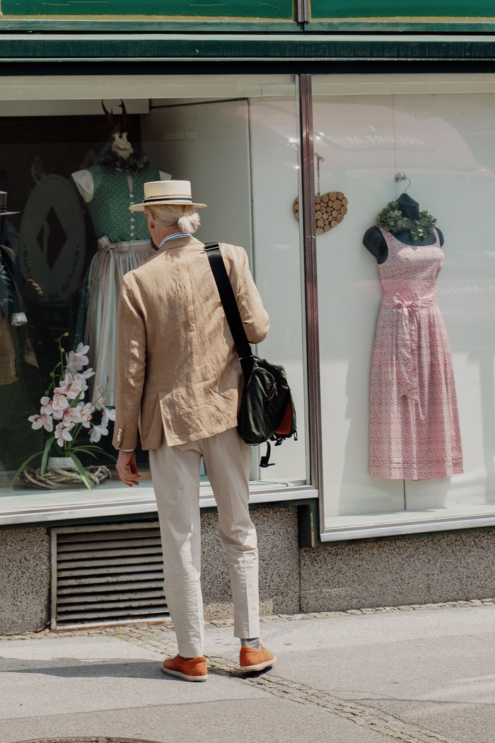 About A stylish man in a hat observes fashion dresses in a store window display.